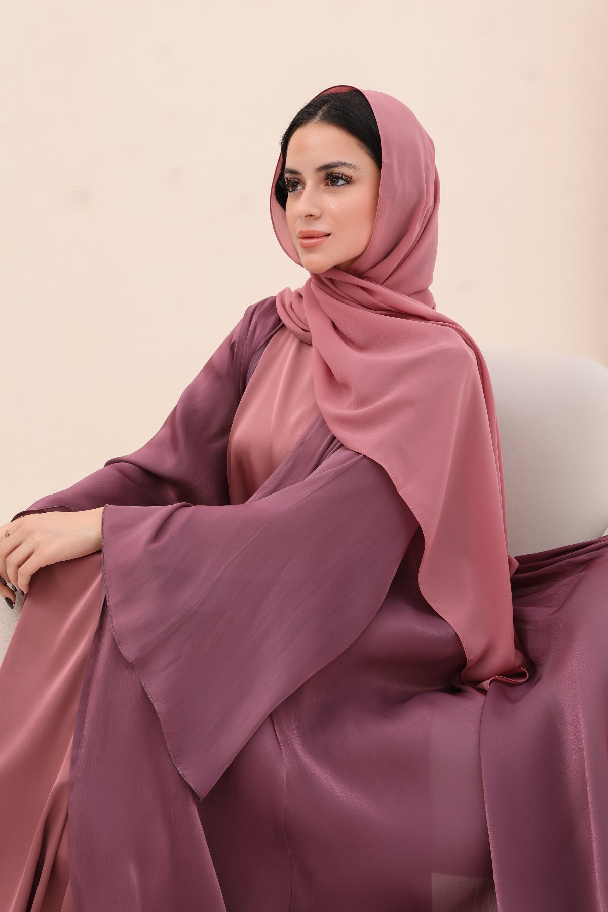 Woman wearing a pink hijab and dress sitting on a chair against a beige background