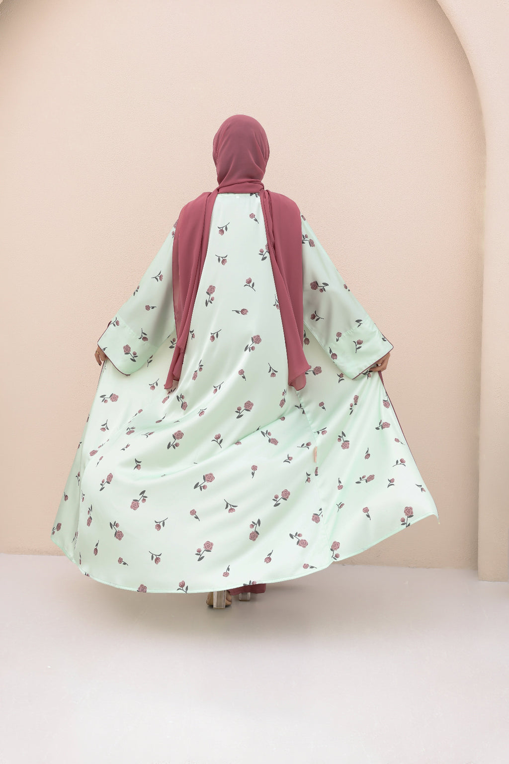 Person wearing a light green dress with floral pattern and a pink hijab on a beige background