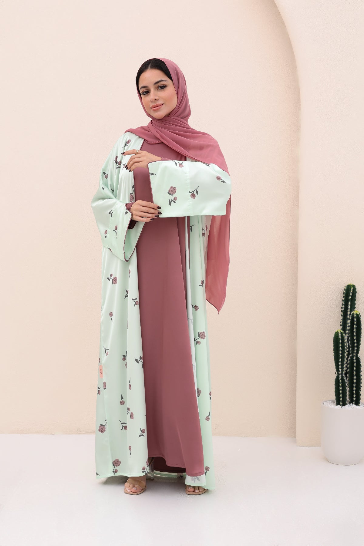 Woman wearing a pink hijab and floral dress holding a clutch against a beige background