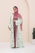 Woman wearing a light green floral abaya and pink hijab against a beige background