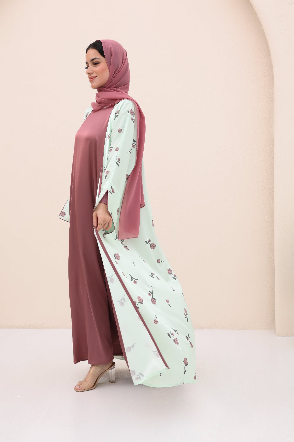 Woman wearing a long, light-colored dress with floral patterns and a matching hijab against a beige background.