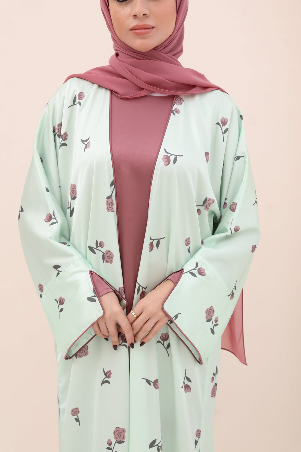 Woman wearing a light green abaya with floral patterns and a pink hijab on a beige background