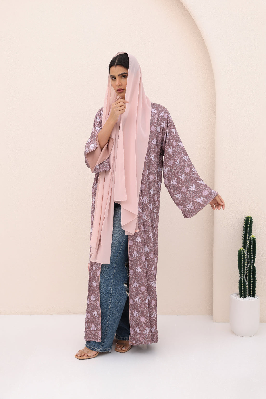 Woman wearing a long floral dress with a pink hijab and blue jeans, standing against a plain background.
