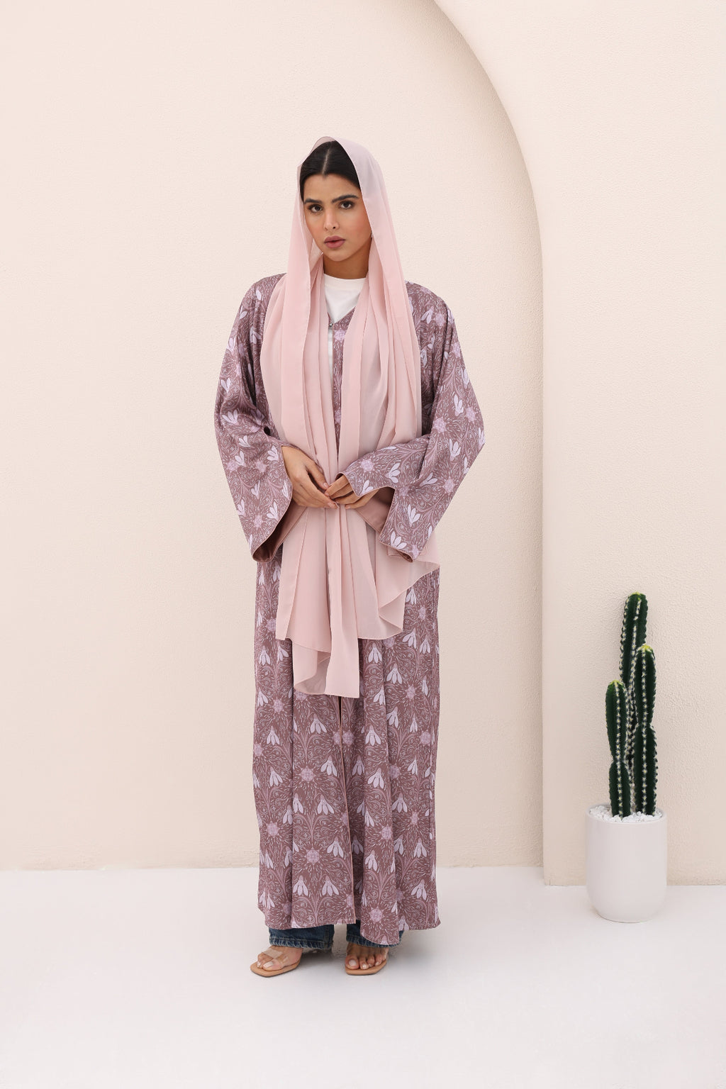 Woman wearing a long, patterned dress with a pink hijab in a minimalistic setting.
