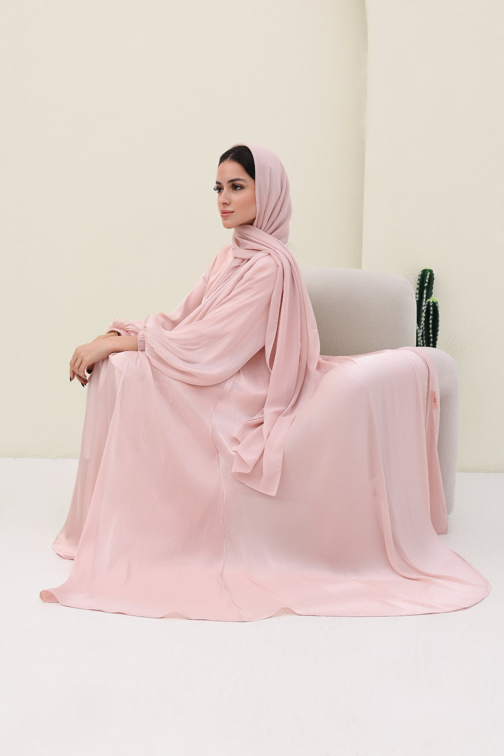 Woman in a pink abaya sitting on a white chair against a light beige wall.