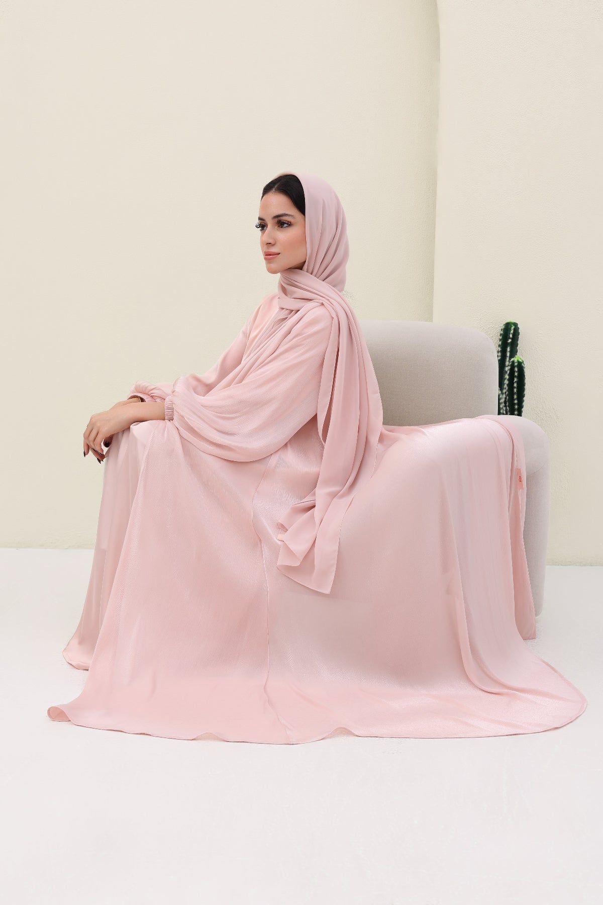 Woman in a pink abaya sitting on a white chair against a light beige wall.