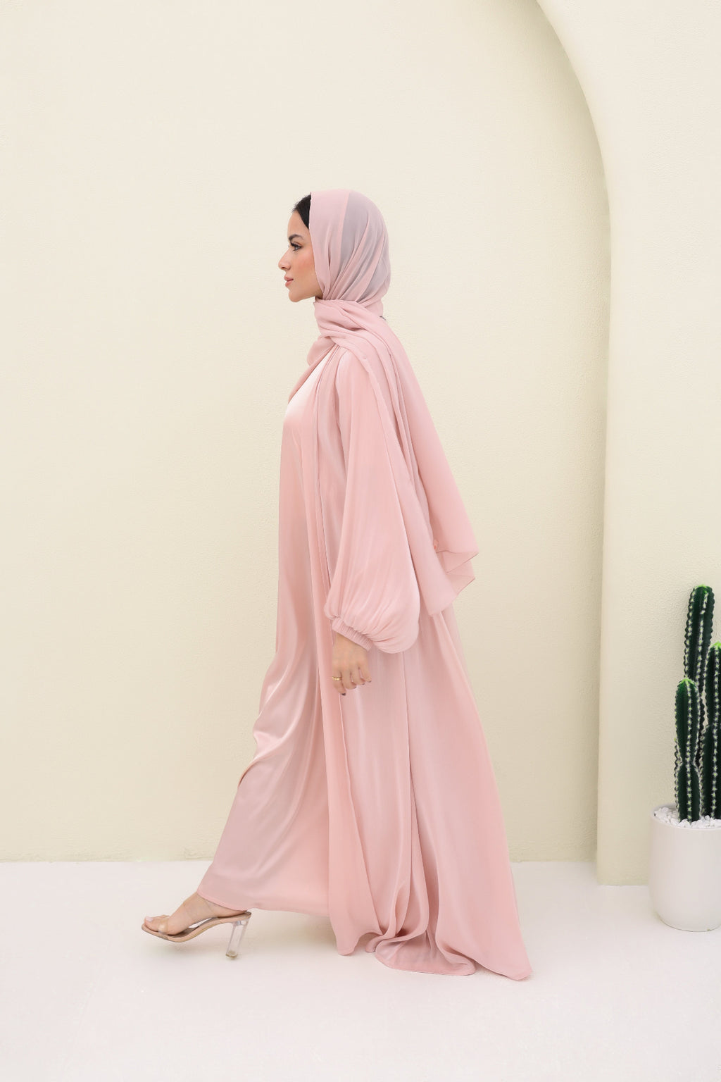 Woman wearing a pink abaya and hijab in a minimalistic setting with a cactus plant.