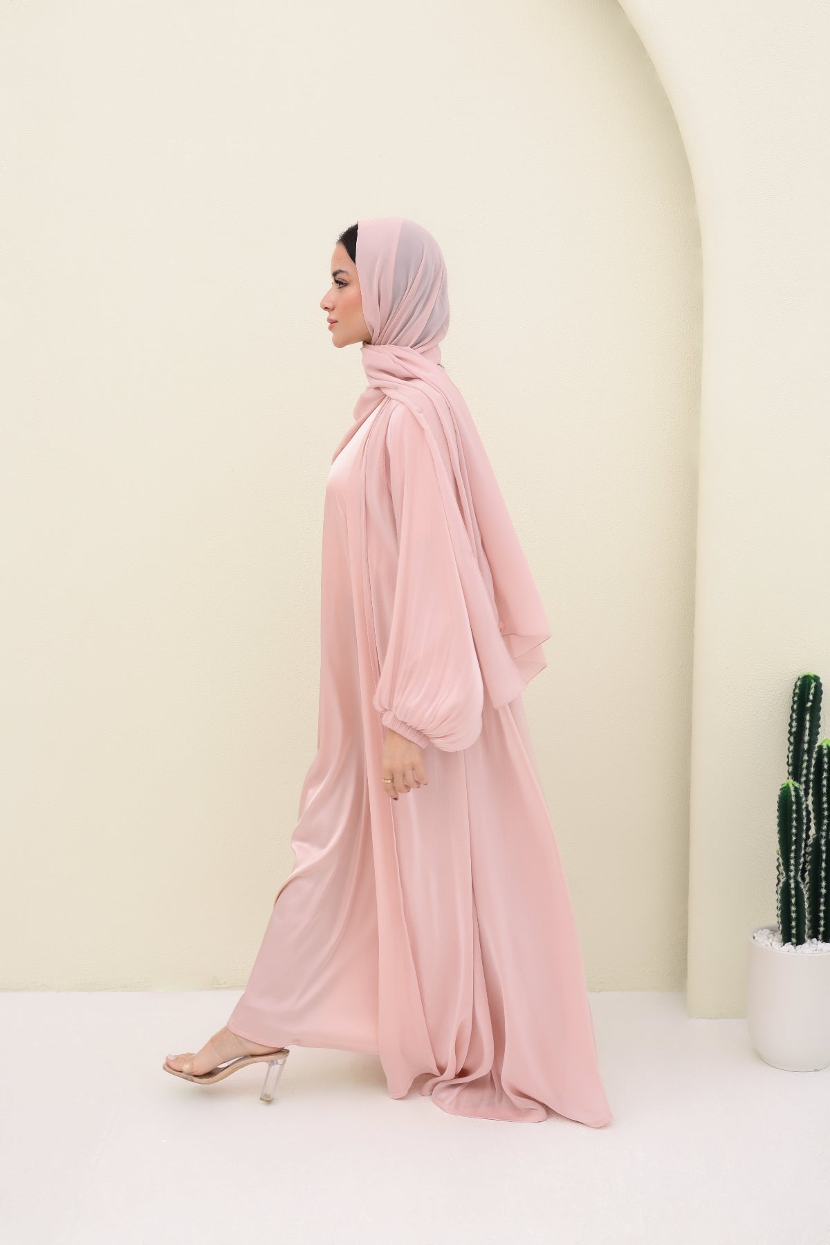 Woman wearing a pink abaya and hijab in a minimalistic setting with a cactus plant.
