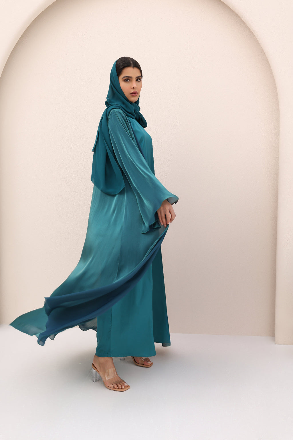 Woman wearing a teal abaya with a matching hijab against a beige background