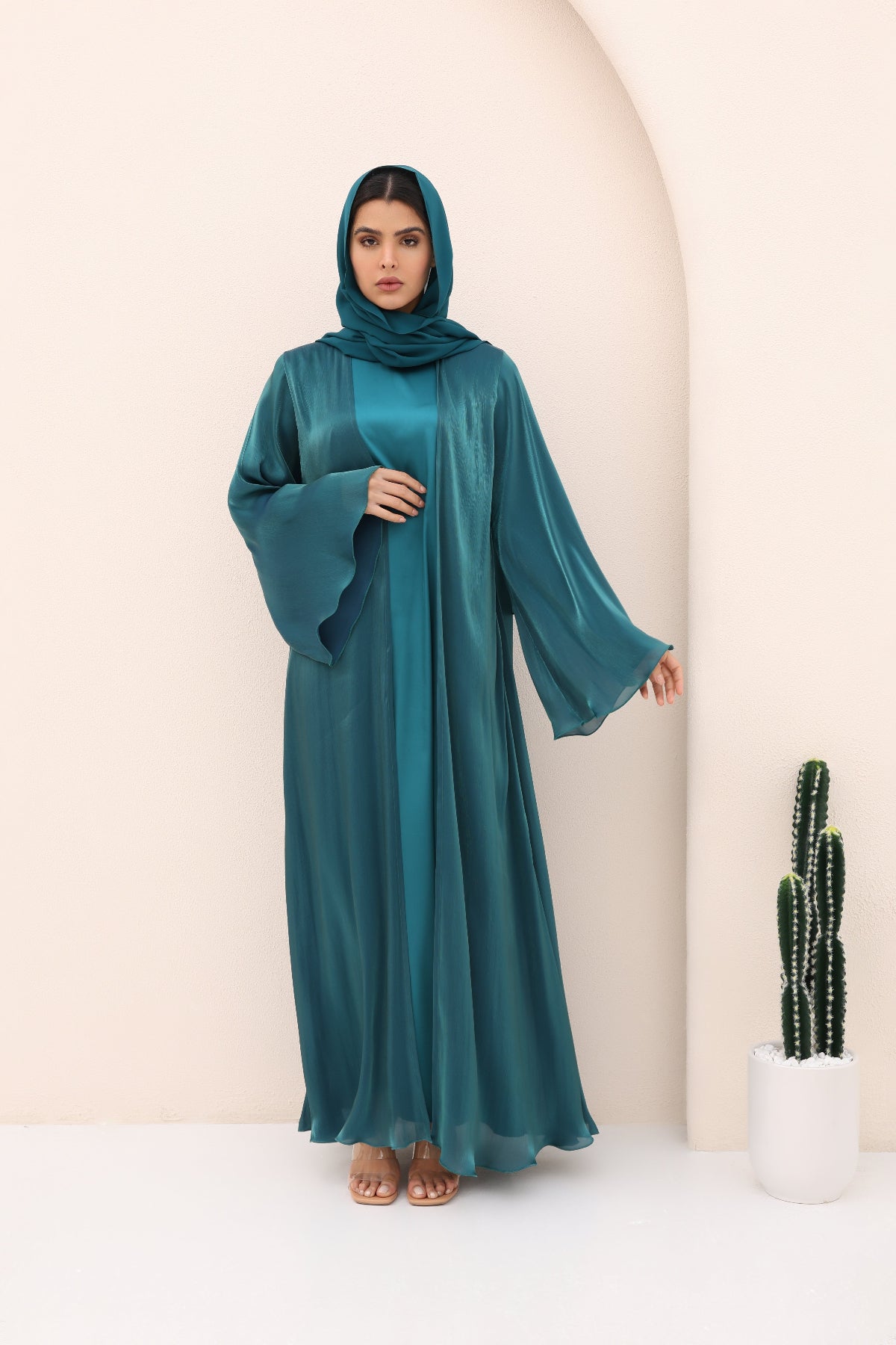 Woman wearing a teal abaya in a minimalistic setting with a cactus plant.