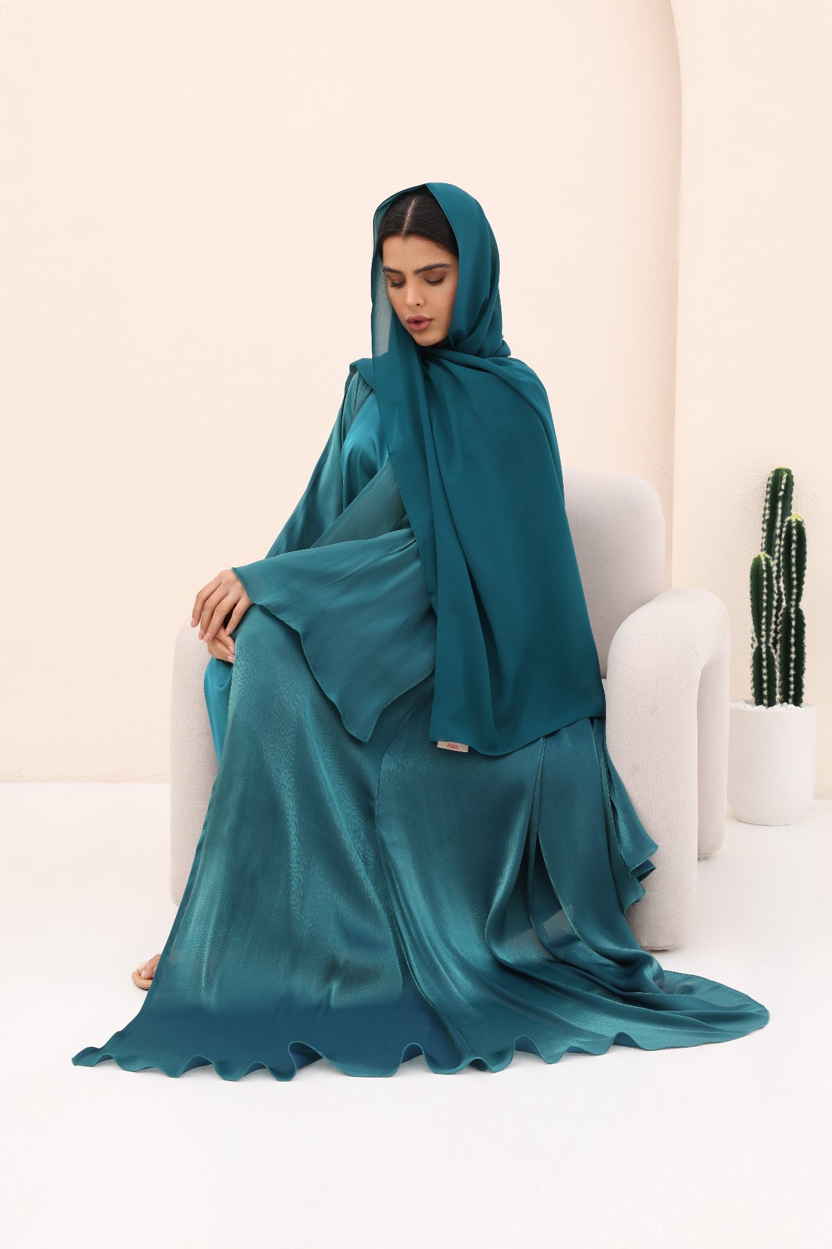 Woman in a teal abaya sitting on a white chair against a beige wall.
