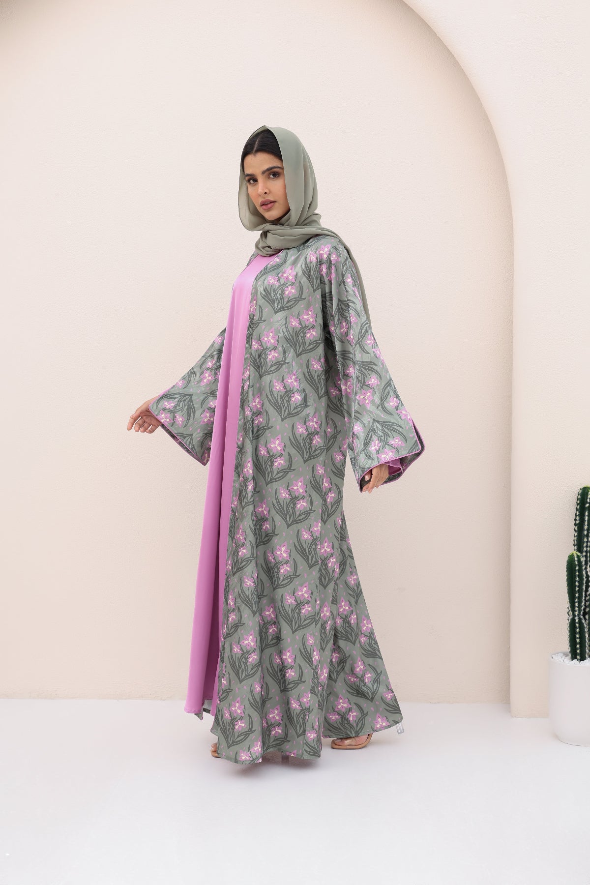 Woman wearing a floral dress with a pink scarf and hijab in a minimalistic setting.